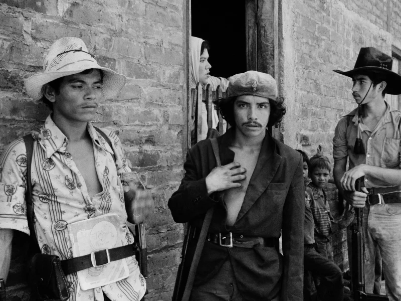 Four men, some armed and in casual clothing, stand near a brick wall in El Salvador as a woman looks out from a doorway and two children stand nearby.