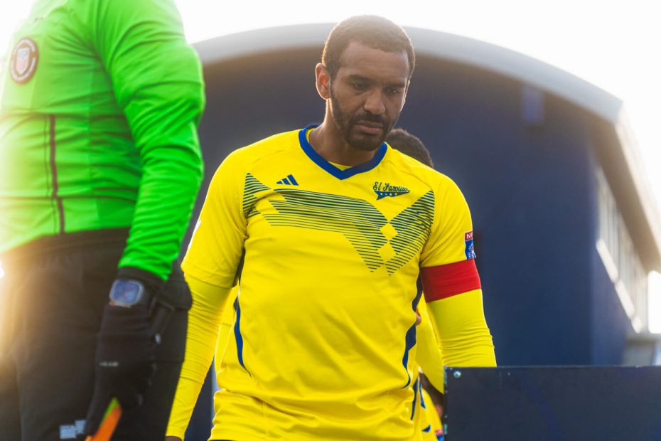 A male soccer player in a yellow uniform with a red captain's armband walks alongside a referee in a green jersey.