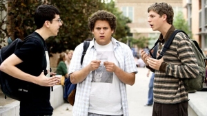 Three teenage boys with backpacks stand outdoors in conversation, two gesturing while the one in the middle looks surprised and holds papers.