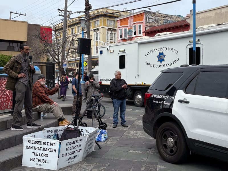 A man speaks with a microphone near police vehicles on a city street. Signs on boxes contain religious messages. Several people stand nearby, and buildings are visible in the background.