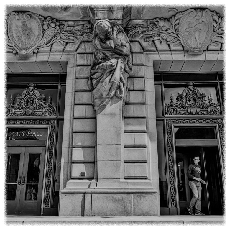 Black and white photo of a building entrance with ornate carvings, a large stone statue above the door, and a person walking out of the doorway on the right.