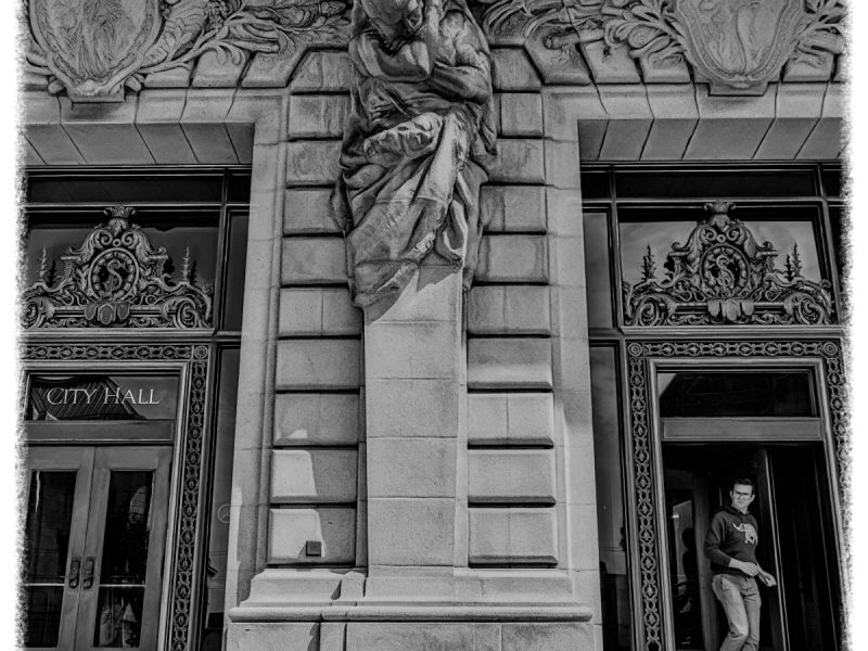 Black and white photo of a building entrance with ornate carvings, a large stone statue above the door, and a person walking out of the doorway on the right.