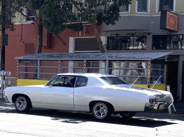 A white classic car is parked on a city street in front of a sidewalk dining area with a yellow barrier and metal roof.