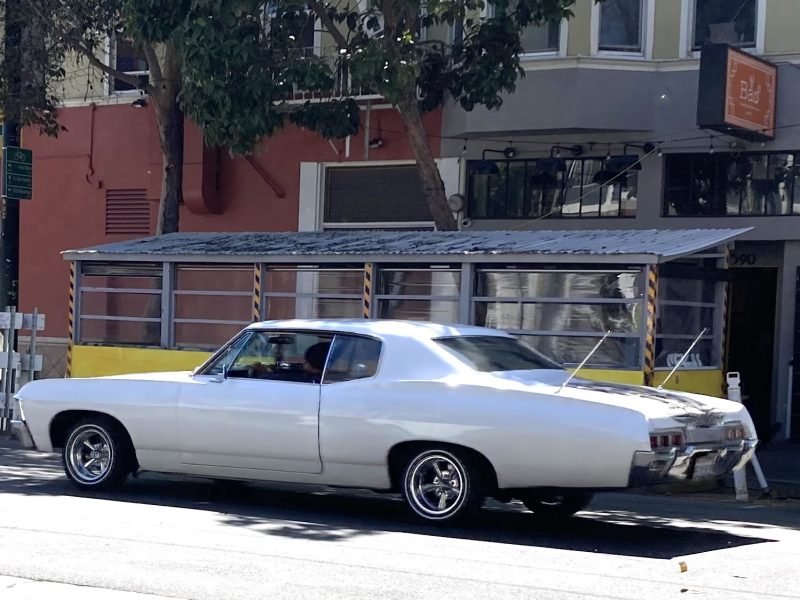 A white classic car is parked on a city street in front of a sidewalk dining area with a yellow barrier and metal roof.
