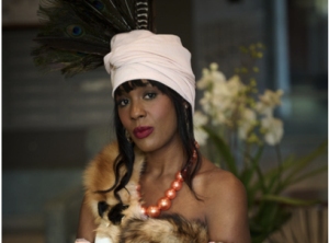 A person wearing a white turban with feather accents, a fur stole, and a large orange bead necklace, stands indoors near a plant with white flowers.
