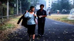 Two people walk side by side on a paved path outdoors, one holding a jacket and the other holding a book, with trees and buildings in the background.
