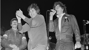 Three men stand on stage; one man claps, another speaks into a microphone, and the third stands beside them, all under stage lighting.