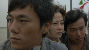 Three people, two men and one woman, sit close together outdoors, looking in different directions with serious expressions.