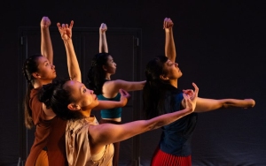 Four dancers perform on stage, each striking different expressive poses with their arms raised against a dark background.