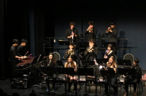 A jazz band of thirteen musicians performs on stage with brass, woodwind, and percussion instruments, all dressed in black attire.