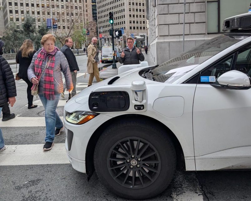 A white self-driving car is stopped at a pedestrian crosswalk next to a busy sidewalk. People are walking in various directions on a city street.