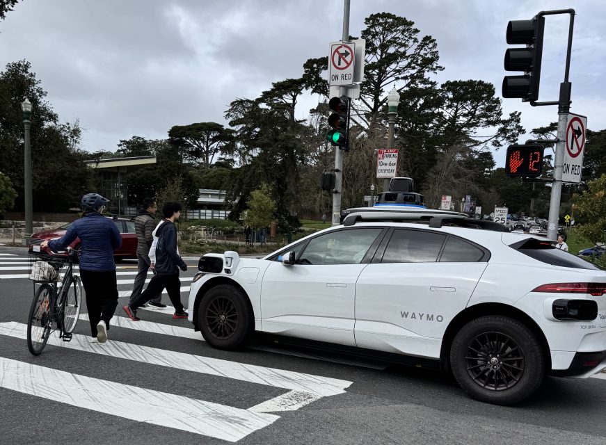 A white autonomous vehicle waits at a crosswalk while a cyclist and pedestrians cross the street. Traffic lights and "No Turn on Red" signs are visible.