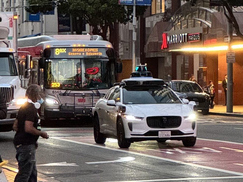 White autonomous vehicle with roof sensors drives in city traffic near a Marriott hotel. A Bayshore Express bus is seen in the background. People walking nearby.