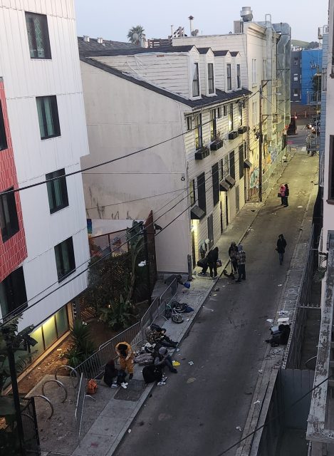 Amidst the 16th-century architecture, an alleyway bustles with people sitting and standing, surrounded by scattered items. Historic buildings line both sides, and a few pedestrians wander through the charming scene.