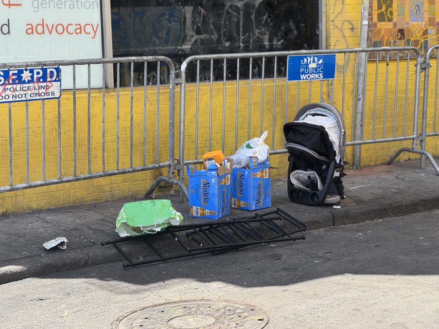 A sidewalk scene shows a stroller, cardboard boxes, a broken metal rack, and scattered items in front of a yellow wall and metal barricades.