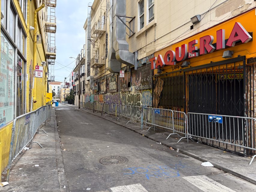 A narrow urban alley lined with buildings and graffiti, metal barricades block the entrance to a closed taqueria with a red sign on the right side.