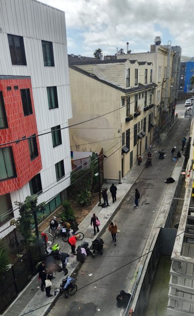 A narrow city alley with groups of people standing, sitting, and pushing strollers near buildings on both sides under a cloudy sky.