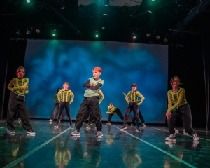Dancers in green and black outfits perform on stage, striking dynamic poses. A blue-lit backdrop and overhead lighting enhance the scene.