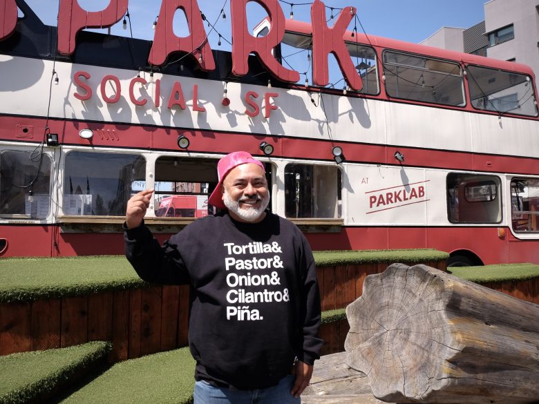A man wearing a black sweatshirt with taco ingredients stands outdoors in front of a red double-decker bus labeled "PARK SOCIAL SF.