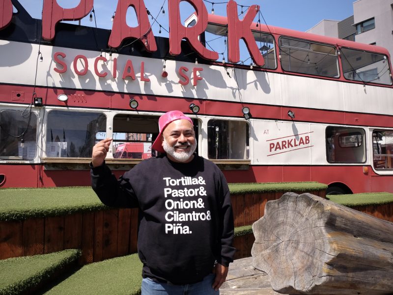 A man wearing a black sweatshirt with taco ingredients stands outdoors in front of a red double-decker bus labeled "PARK SOCIAL SF.