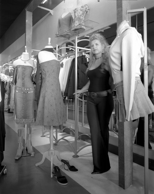 A woman stands in a clothing store next to mannequins dressed in various outfits, with racks of clothes and footwear visible in the background.