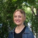 A person with blonde hair smiles while standing in front of green foliage, wearing a black top and denim jacket.