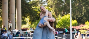 Two dancers in blue costumes perform a contemporary dance outdoors, with trees and columns in the background and people watching nearby.