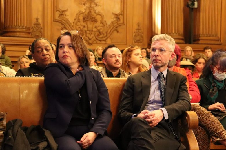 Several people sit attentively in a wood-paneled room, including a woman and a man in business attire in the front row, as the audience listens closely to discussions on budget cuts.