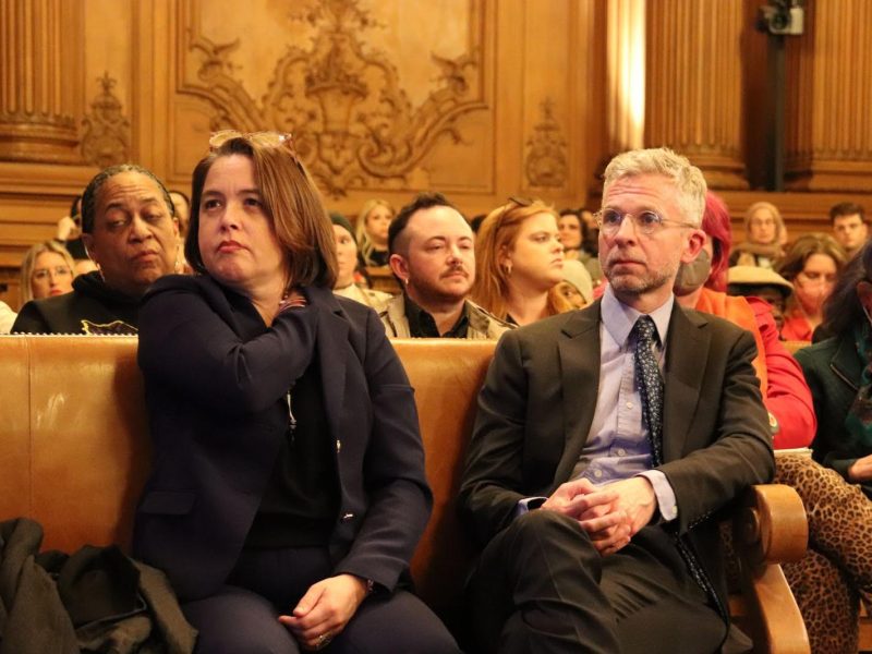 Several people sit attentively in a wood-paneled room, including a woman and a man in business attire in the front row, as the audience listens closely to discussions on budget cuts.