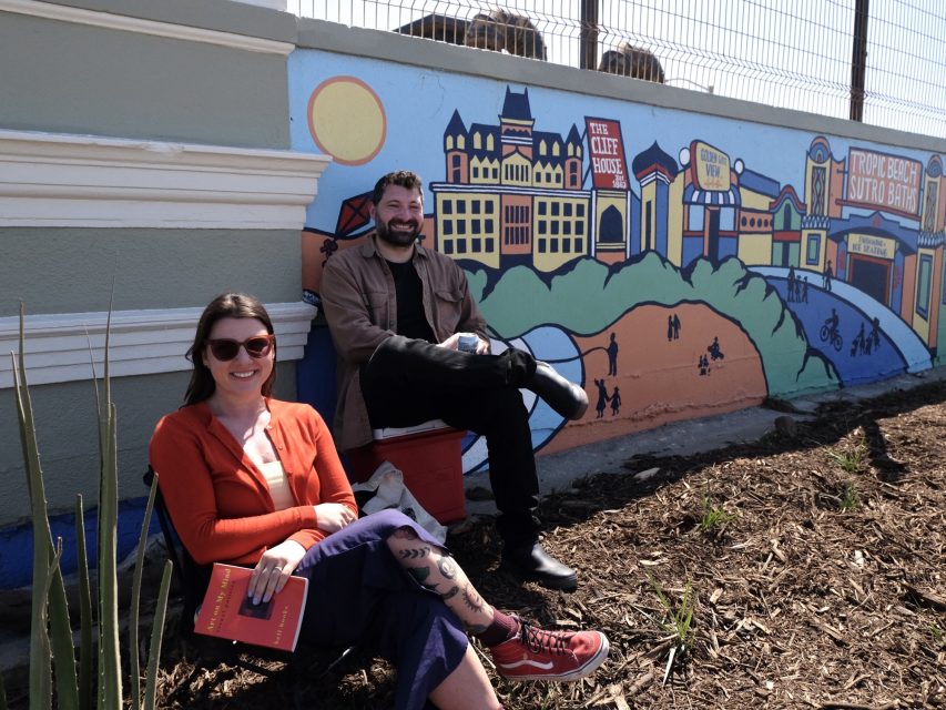 Two people sit in front of a colorful mural depicting a cityscape. The person on the left is holding a book and smiling. The scene includes plants and a fence overhead.