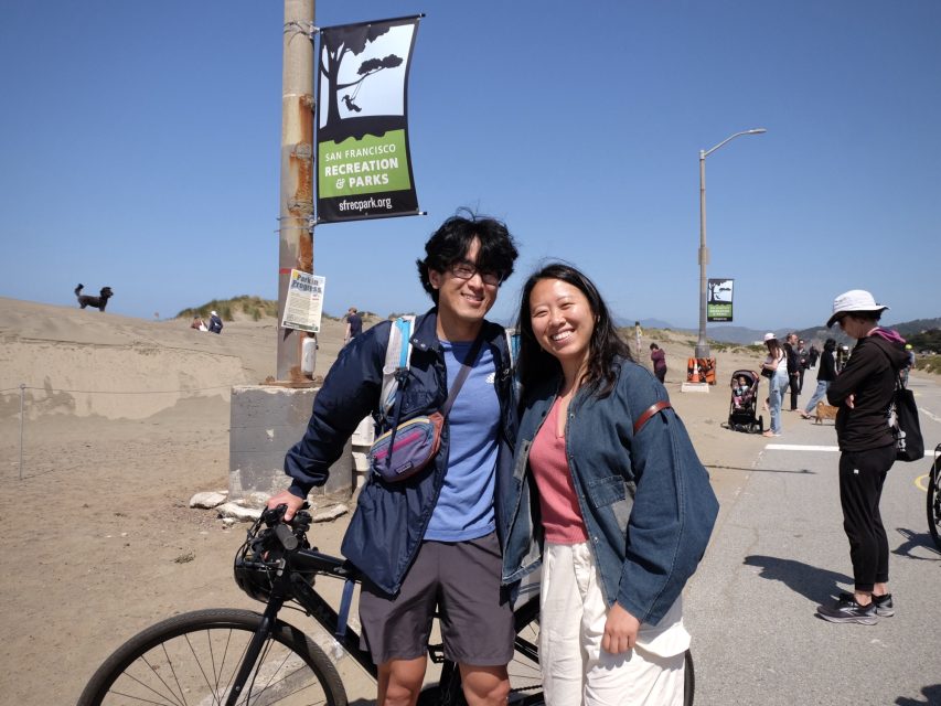 A couple stands smiling beside a bike, with a beach and blue sky in the background. A sign for San Francisco Recreation & Parks is visible nearby.