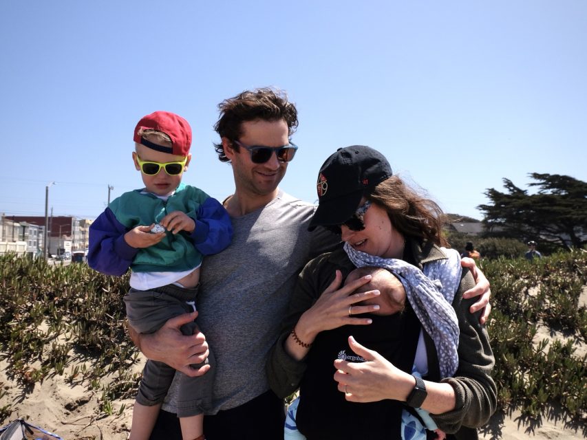 A man and woman stand on a beach, with the man holding a young child in sunglasses and the woman holding a baby in a carrier. The sky is clear and the background shows greenery.