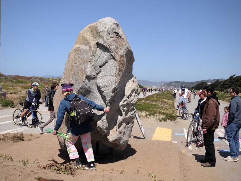 People gather around a large rock on a sandy area near a pathway. Some are cycling, and others are walking. The sky is clear and blue.