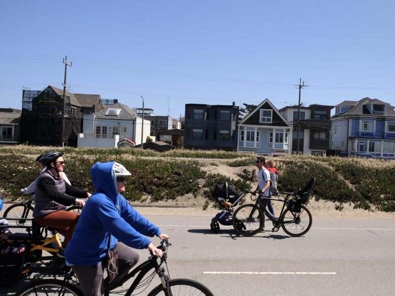 People riding bicycles along a road near residential houses on a clear day. Some cyclists wear helmets. A stroller is attached to one bicycle.