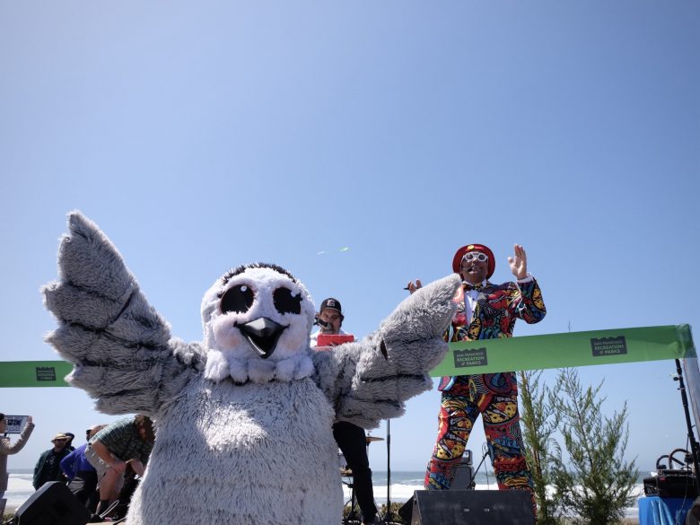 A person in a gray owl costume stands in the foreground, while a person in a vibrant, patterned suit and hat gestures behind a green ribbon on a sunny day.