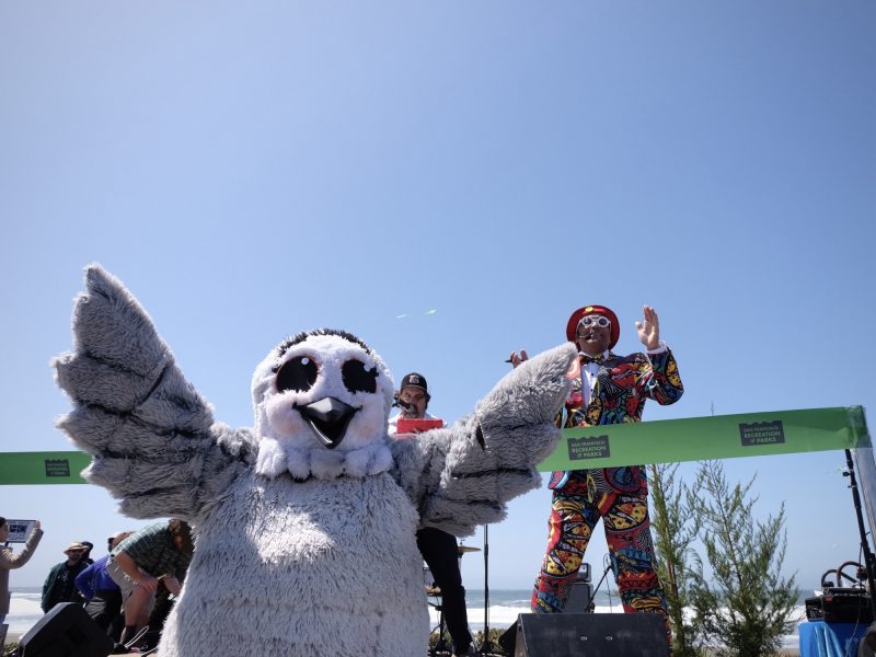 A person in a gray owl costume stands in the foreground, while a person in a vibrant, patterned suit and hat gestures behind a green ribbon on a sunny day.