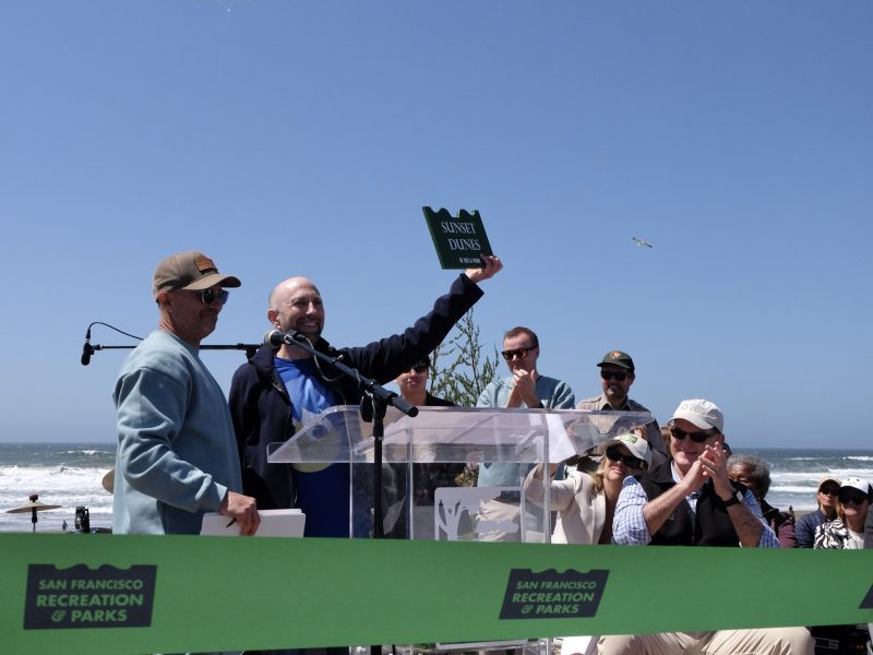 A man holds up a "Sully Davis" street sign at an outdoor event near the ocean, with people seated and standing around a podium and a green San Francisco Recreation & Parks ribbon in front.