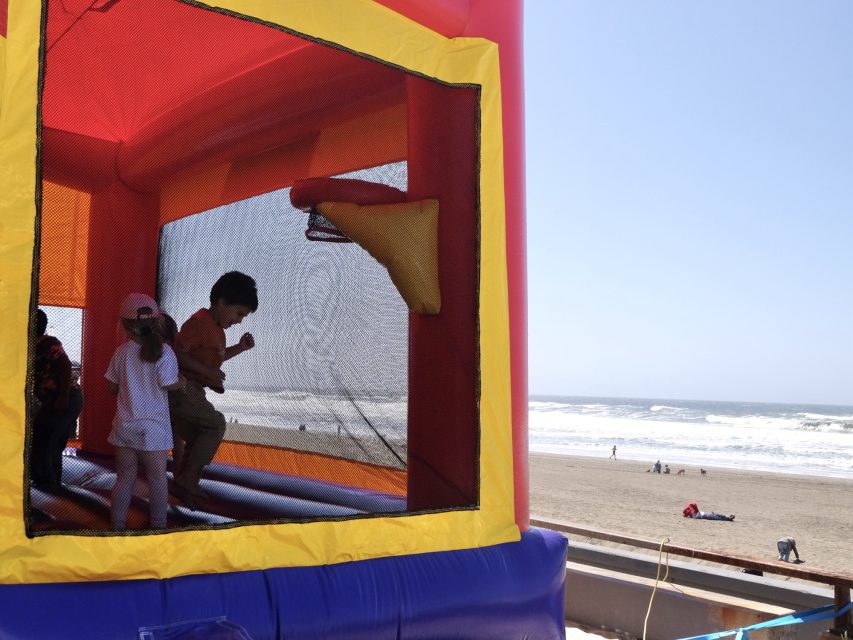Children play inside a colorful inflatable bounce house near a beach with people and waves in the background.