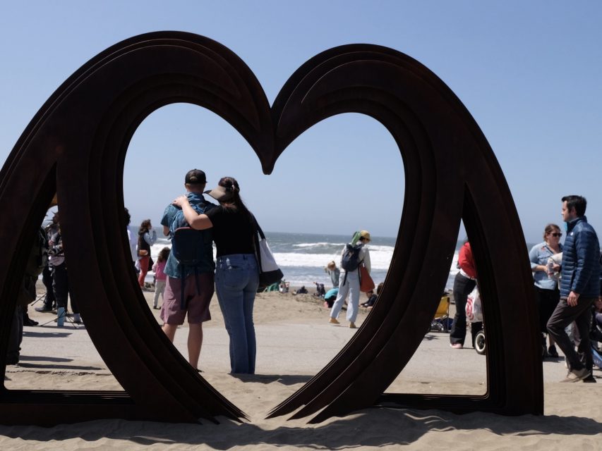 A couple stands embraced, framed by a large heart-shaped sculpture on a busy beach with people and ocean waves in the background.