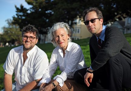 Three adults, two men and one woman, sit on grass outdoors in casual clothing, looking at the camera with houses and trees in the background.
