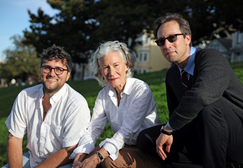Three adults, two men and one woman, sit on grass outdoors in casual clothing, looking at the camera with houses and trees in the background.