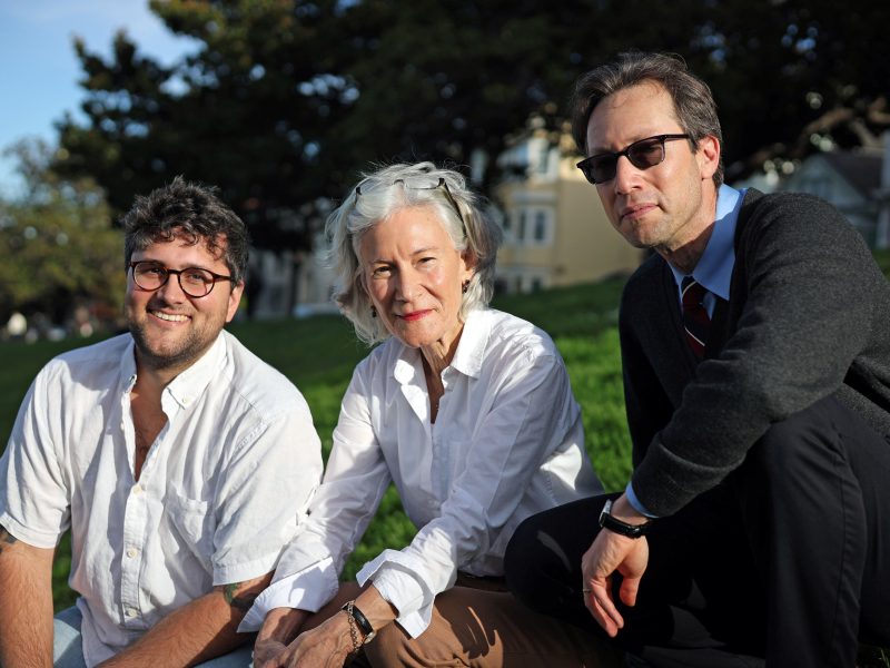 Three adults, two men and one woman, sit on grass outdoors in casual clothing, looking at the camera with houses and trees in the background.