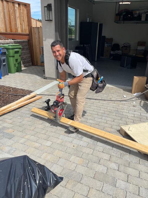 A man using a circular saw to cut a wooden plank outdoors on a paved surface, wearing work gloves and a tool belt, with a garage and various materials in the background.