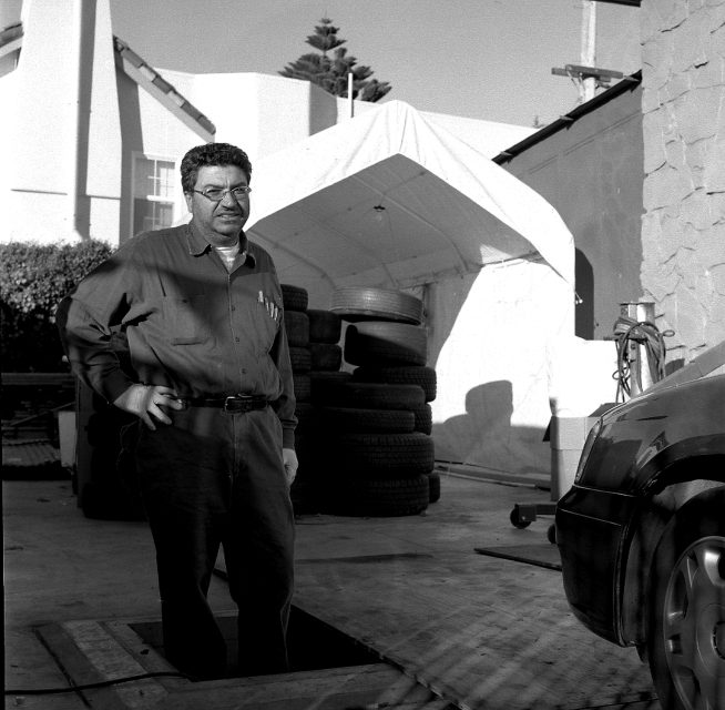 A man stands near a car and a pile of tires outside a building, with a tented structure and shrubbery in the background.