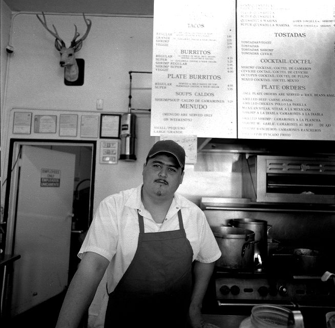A man in a white shirt and apron stands behind the counter of a restaurant kitchen, with a menu board and mounted deer head visible in the background.