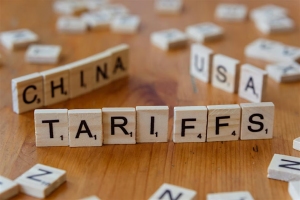 Scrabble tiles on a wooden surface spell out "TARIFFS," with "CHINA" and "USA" also formed from tiles in the background.