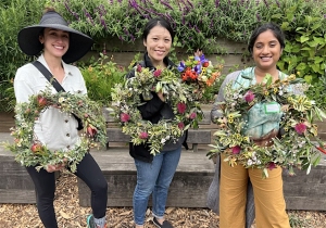 Three women stand outdoors holding handmade floral wreaths, smiling at the camera with greenery and flowers in the background.