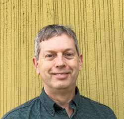 A person with short gray hair and a green shirt is smiling in front of a textured yellow wall.