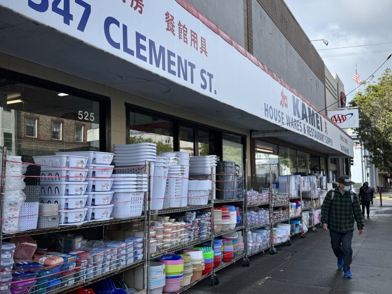 A storefront displaying household items in bins and shelves, including plastic containers and colorful bowls. A person walks on the sidewalk in front of the store.