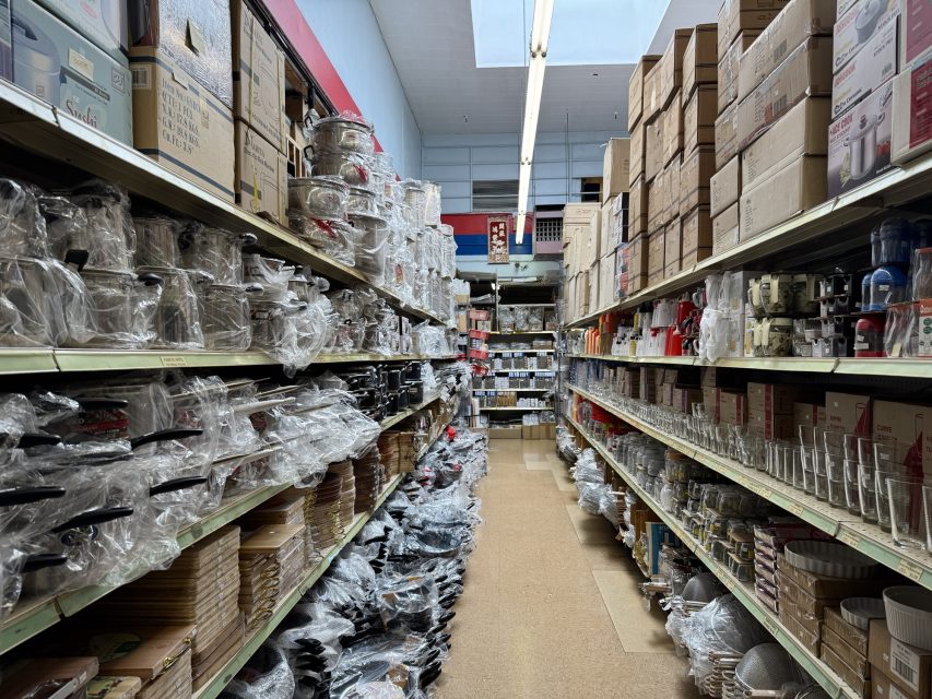 A store aisle filled with stacked kitchenware and appliances, like pans and glassware, on both sides. Boxes are stacked high on the upper shelves.